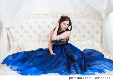 Attractive brunette woman with beautiful makeup in blue evening dress posing while sitting on bed, touching her hairs and looking at camera with pasion. indoor studio shot. Attractive brunette woman with beautiful makeup in blue evening dress posing while sitting on bed, touching her hairs and looking at camera with pasion. indoor studio shot. 89354843