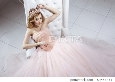 Top view portrait of beautiful young woman in crown and luxurious pink dress posing on stylish white armchair, sitting and looking at camera. indoor studio shot. 89354855