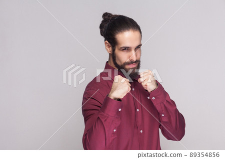 Portrait of serious handsome man with dark collected long hair and beard in red shirt standing, looking at camera with boxing fists and ready to attack. indoor studio shot, isolated on gray background 89354856