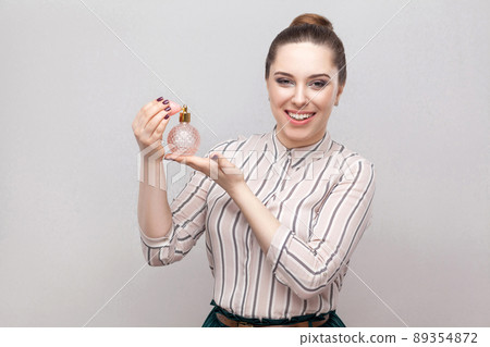 Woman using perfume. Smiling attractive woman spraying perfume enjoying aroma fragrance holding bottle of perfume isolated gray background. Indoor, studio sho, isolated on gray background, copy space 89354872