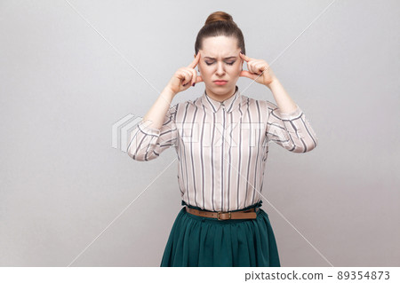 Headache. Portrait of unhappy beautiful young woman in striped shirt and green skirt and collected ban hairstyle, standing and holding her painful head. indoor studio shot, isolated on grey background Headache. Portrait of unhappy beautiful young woman in striped shirt and green skirt and collected ban hairstyle, standing and holding her painful head. indoor studio shot, isolated on grey background 89354873