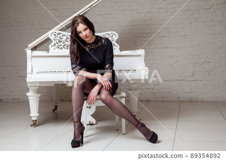 Sensual brunette woman in lace black classic dress sitting near white piano and looking at camera. indoor studio shot on white brick wall. Sensual brunette woman in lace black classic dress sitting near white piano and looking at camera. indoor studio shot on white brick wall. 89354892