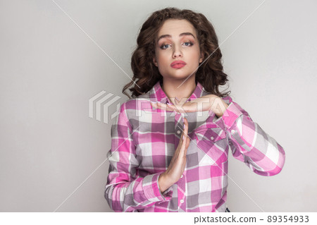 Please give me more time. Satisfied beautiful girl with pink checkered shirt, standing and looking at camera and pleading for timeout or pause . indoor studio shot, isolated on gray background. 89354933