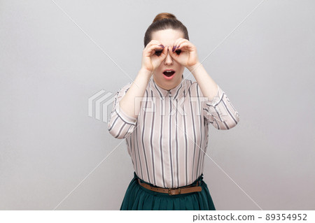 Portrait of funny beautiful young woman in striped shirt and green skirt with makeup and collected ban hairstyle, standing and looking with binoculars gesture. Studio shot, isolated on grey background Portrait of funny beautiful young woman in striped shirt and green skirt with makeup and collected ban hairstyle, standing and looking with binoculars gesture. Studio shot, isolated on grey background 89354952