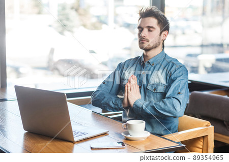 Time to relax! Portrait of handsome successful bearded young freelancer in blue jeans shirt are sitting in cafe and having a rest, holding hands like have meditative to have emotional pleasure. indoor 89354965