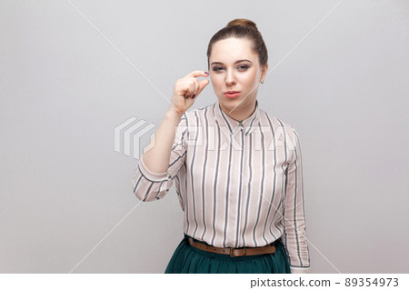 Give me a little bit more please. Portrait of hopeful beautiful young woman in striped shirt with makeup and collected ban hairstyle, standing and pleading. studio shot, isolated on grey background. 89354973