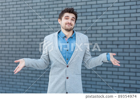 I don't know. Portrait of confused handsome bearded man in casual style standing, raised arms and looking at camera with doubtful face. indoor studio shot on brick wall background. 89354974
