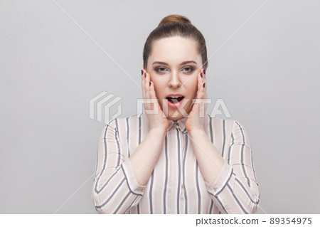 Portrait of happy surprised beautiful young woman in striped shirt, collected ban hairstyle, standing, amazed and looking at camera with toothy smile. indoor studio shot, isolated on grey background. Portrait of happy surprised beautiful young woman in striped shirt, collected ban hairstyle, standing, amazed and looking at camera with toothy smile. indoor studio shot, isolated on grey background. 89354975