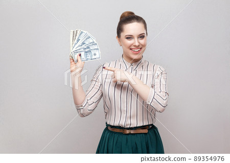 Happy satisfied attractive young businesswoman in striped shirt standing, holding many dollars, pointing finger and looking at camera with toothy smile. Indoor, studio shot, isolated, gray background 89354976