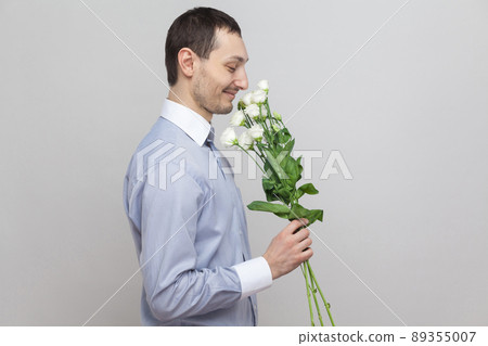 He wants to give a flowers. Side view portrait of charming pleasure romantic young man in blue shirt holding bouquet of white flowers smelling them. Indoor, studio shot, isolated on gray background 89355007