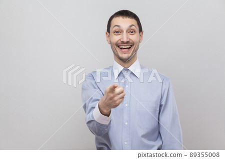 Is that was you? Amazed handsome bristle businessman in classic blue shirt standing, pointing and looking at camera with surprised face. indoor studio shot, isolated on grey background copyspace. 89355008