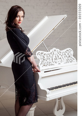 Side view profile of sensual brunette woman in lace black classic dress standing near white piano and looking at camera. indoor studio shot on white brick wall. 89355009