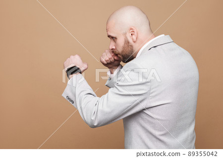 Profile side view portrait of middle aged bald bearded businessman in light gray suit standing and ready to attack with boxer pose. indoor studio shot, isolated on light brown background. 89355042