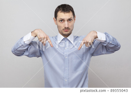 Here and right know. Portrait of serious handsome bristle businessman in classic blue shirt standing and looking at camera and pointing down. indoor studio shot, isolated on grey background copyspace. 89355043