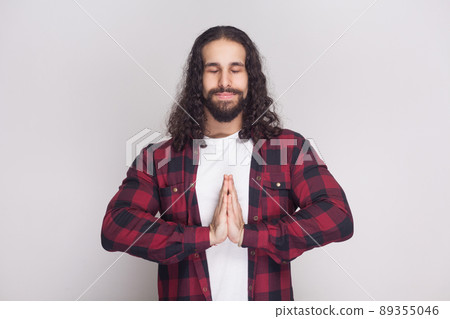 Portrait of calm meditative handsome man with beard and black long curly hair in casual style, checkered red shirt standing with clossed eyes and yoga gesture pose. indoor isolated on grey background. 89355046