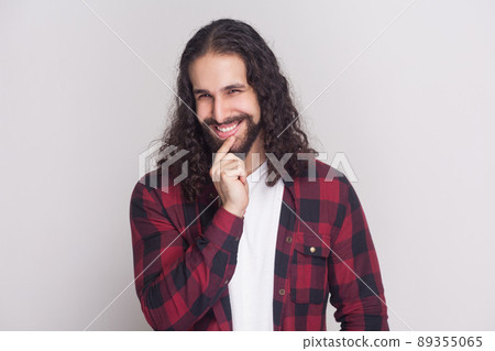 Portrait of playful handsome man with beard and black long curly hair in casual style, checkered red shirt standing looking at camera with cunning face. indoor studio shot, isolated on grey background 89355065