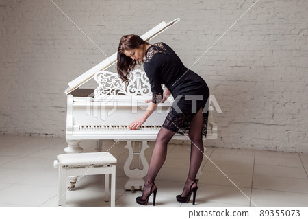 Profile side view of seductive brunette woman in dark classic dress standing playing piano. indoor studio shot on white brick wall. 89355073