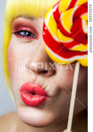 Beauty portrait of funny cute young female model with freckles, red makeup and yellow wig, holding colorful candy stick, kissing and looking at camera. indoor studio shot, isolated on gray background. Beauty portrait of funny cute young female model with freckles, red makeup and yellow wig, holding colorful candy stick, kissing and looking at camera. indoor studio shot, isolated on gray background. 89355074