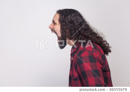 Profile side view portrait of handsome angry man with beard and black long curly hair in casual style, checkered red shirt standing and screaming. indoor studio shot, isolated on grey background. 89355079
