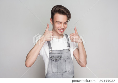 Happy young brunette man in casual style with white t-shirt and denim overalls standing and looking at camera with toothy smile and thumbs up. indoor studio shot, isolated on gray background. Happy young brunette man in casual style with white t-shirt and denim overalls standing and looking at camera with toothy smile and thumbs up. indoor studio shot, isolated on gray background. 89355082