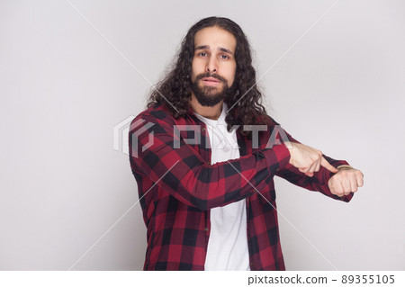 we have no more time. serious man with beard and black long curly hair in casual style, checkered red shirt standing showing time gesture and worry. indoor studio shot, isolated on grey background. we have no more time. serious man with beard and black long curly hair in casual style, checkered red shirt standing showing time gesture and worry. indoor studio shot, isolated on grey background. 89355105