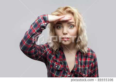Portrait of surprised beautiful blonde young woman in casual red checkered shirt standing and looking at camera with hand on forhead. indoor studio shot, isolated on grey background. 89355106