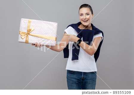 Beautiful wondered woman pointing present for you gift box with bow with blue jeans and sweater on her shoulders and smart watch, Expression and feelings concept. indoor, isolated on gray background 89355109