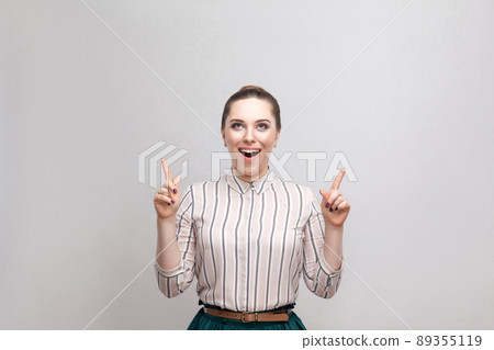 Surprised beautiful young woman in striped shirt and green skirt with makeup and collected ban hairstyle, standing with amazed face and pointing at copyspace. studio shot, isolated on grey background. Surprised beautiful young woman in striped shirt and green skirt with makeup and collected ban hairstyle, standing with amazed face and pointing at copyspace. studio shot, isolated on grey background. 89355119