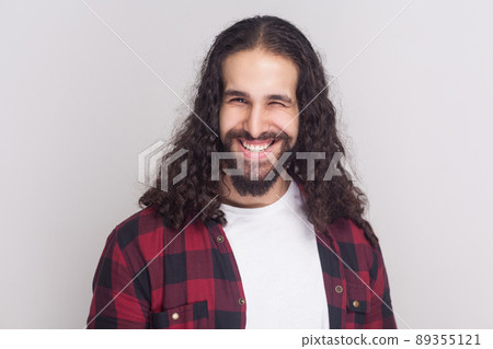 Funny handsome man with beard and black long curly hair in casual style, checkered red shirt standing, winking and looking at camera with toothy smile. indoor studio shot, isolated on grey background 89355121