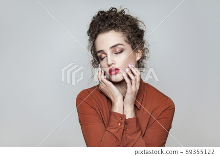 Beauty portrait of young beautiful model with collected dark curly hair, nude makeup, red lips, and brown shirt touching her face with closed eys. indoor studio shot. isolated on gray background. Beauty portrait of young beautiful model with collected dark curly hair, nude makeup, red lips, and brown shirt touching her face with closed eys. indoor studio shot. isolated on gray background. 89355122
