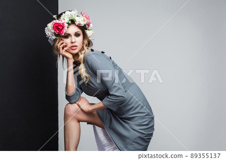 Portrait of beautiful sensual fashion model in grey dress with makeup and head flowers posing and looking at camera and touching her face. indoor studio shot, isolated on black and grey background. 89355137