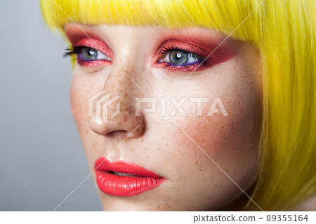 Closeup beauty portrait of calm cute young female model with freckles, red makeup and yellow wig, looking forward with serious face. indoor studio shot, isolated on gray background. Closeup beauty portrait of calm cute young female model with freckles, red makeup and yellow wig, looking forward with serious face. indoor studio shot, isolated on gray background. 89355164