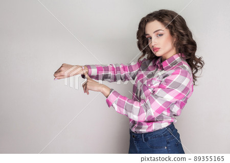 Time is gone. serious beautiful girl with pink checkered shirt, curly hairstyle and makeup standing showing time gesture and looking at camera. indoor studio shot, isolated on gray background. Time is gone. serious beautiful girl with pink checkered shirt, curly hairstyle and makeup standing showing time gesture and looking at camera. indoor studio shot, isolated on gray background. 89355165
