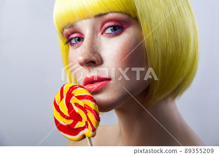 Beauty portrait of cute young female model with freckles, red makeup and yellow wig, holding colorful candy stick and looking at camera, serious face. indoor studio shot, isolated on gray background. 89355209
