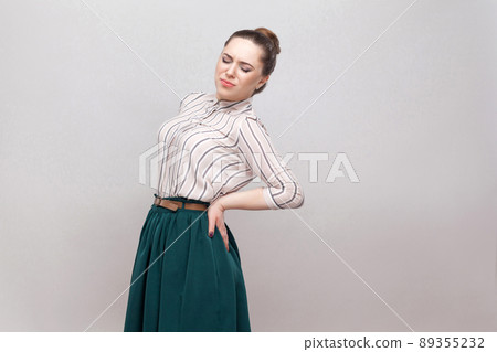 Spine or kidney pain. Portrait of beautiful young woman in striped shirt and green skirt and collected ban hairstyle, standing and holding her painful back. studio shot, isolated on grey background. Spine or kidney pain. Portrait of beautiful young woman in striped shirt and green skirt and collected ban hairstyle, standing and holding her painful back. studio shot, isolated on grey background. 89355232