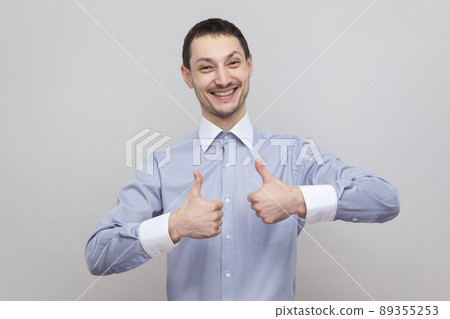 Portrait of satisfied handsome bristle businessman in classic light blue shirt standing and looking at camera with toothy smile and thumbs up. indoor studio shot, isolated on grey background copyspace Portrait of satisfied handsome bristle businessman in classic light blue shirt standing and looking at camera with toothy smile and thumbs up. indoor studio shot, isolated on grey background copyspace 89355253