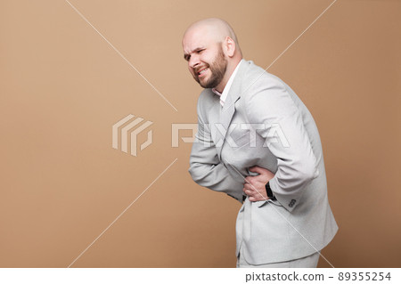 Stomach pain. profile side view portrait of middle aged bald bearded businessman in light gray suit standing and holding his painful belly. indoor studio shot, isolated on light brown background. 89355254