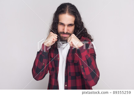 Portrait of serious handsome man with beard and black long curly hair in casual checkered red shirt standing, looking at camera and ready to attack. indoor studio shot, isolated on grey background. 89355259