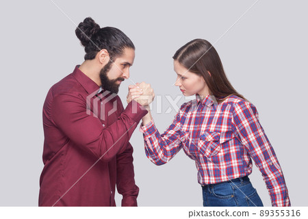 Profile side view portrait of bearded man and woman in casual style standing in armwrestling pose and looking at each other with serious face. indoor studio shot, isolated on gray background. 89355316