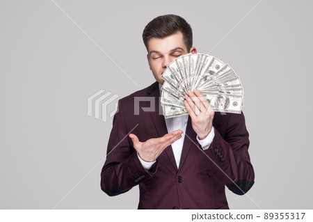 Smell of richness. Satisfied attractive young businessman in classic violet suit standing, holding money and proud with closed eyes and smelling. Indoor studio shot, isolated, gray background. 89355317