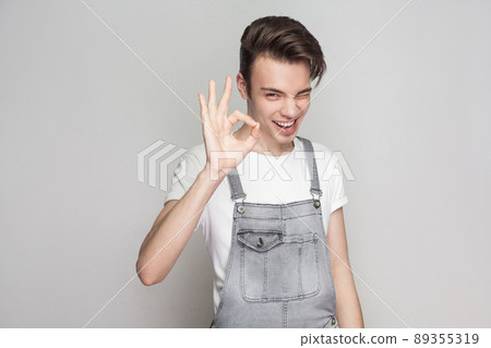 Happy young brunette man in casual style with t-shirt and denim overalls standing and looking at camera with toothy smile, winking and showing Ok sign. indoor studio shot, isolated on gray background 89355319