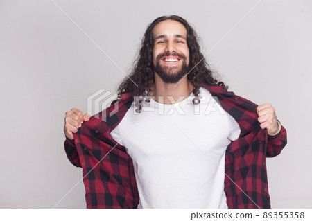 Happy toothy smile handsome man with beard and black long curly hair in checkered standing looking at camera, holding his red shirt and showing white shirt. studio shot, isolated on grey background. Happy toothy smile handsome man with beard and black long curly hair in checkered standing looking at camera, holding his red shirt and showing white shirt. studio shot, isolated on grey background. 89355358
