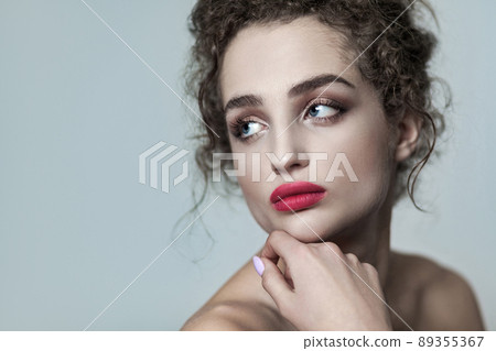 Beauty portrait of young beautiful fashion model with collected dark curly hair, nude makeup and red lips looking away serious with hand on chin. indoor studio shot. isolated on gray background. 89355367