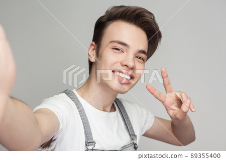 Selfie time! Happy handsome brunette blogger teenager wearing in overalls and white t-shirt standing, showing v sing, making selfie with toothy smile. Indoor, isolated, studio shot, grey background Selfie time! Happy handsome brunette blogger teenager wearing in overalls and white t-shirt standing, showing v sing, making selfie with toothy smile. Indoor, isolated, studio shot, grey background 89355400