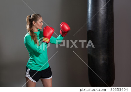 Side view of athlete woman in green long sleeve and black shorts standing in attack position, ready to fight punching bag in boxing red gloves. Indoor studio shot, isolated on dark grey background. 89355404