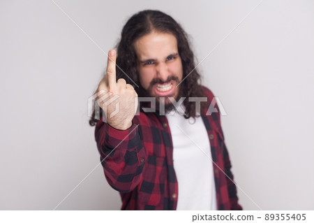 Portrait of angry handsome man with beard and black long curly hair in casual checkered red shirt standing looking at camera and showing middle finger. indoor studio shot, isolated on grey background 89355405
