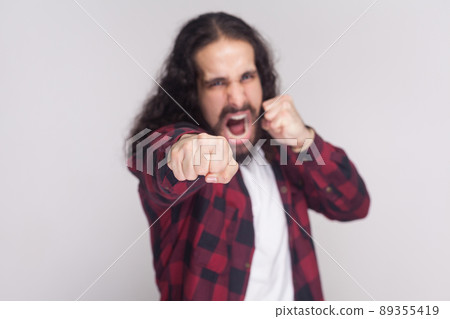 Angry screaming man with beard and black long curly hair in casual checkered red shirt standing, looking at camera and attacking with boxing fists . indoor studio shot, isolated on grey background 89355419