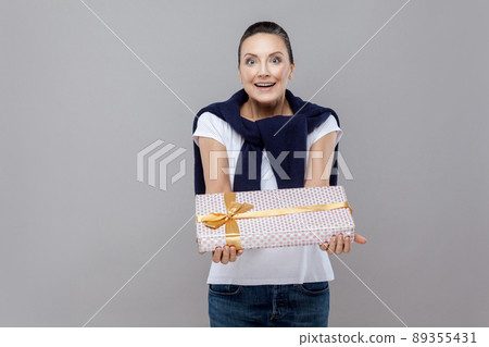 Beautiful woman present for you gift box with bow with blue jeans and sweater on her shoulders. Expression emotion and feelings concept. Studio shot, isolated on gray background Beautiful woman present for you gift box with bow with blue jeans and sweater on her shoulders. Expression emotion and feelings concept. Studio shot, isolated on gray background 89355431
