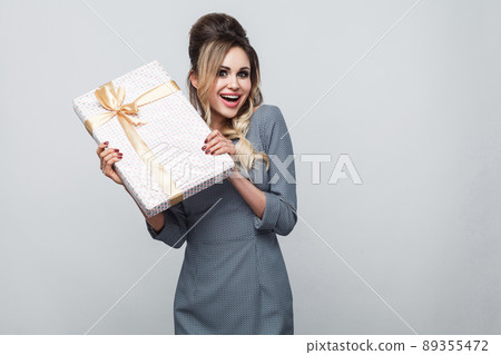Portrait of emotional pleasure beautiful young woman in grey dress standing and showing you present with yellow bow and toothy smile, looking at camera. Indoor, isolated, studio shot, gray background Portrait of emotional pleasure beautiful young woman in grey dress standing and showing you present with yellow bow and toothy smile, looking at camera. Indoor, isolated, studio shot, gray background 89355472