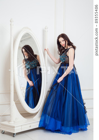 Gorgeous brunette woman in fashionable dress posing near big mirror at white room and looking at camera and smiling. indoor studio shot. 89355486